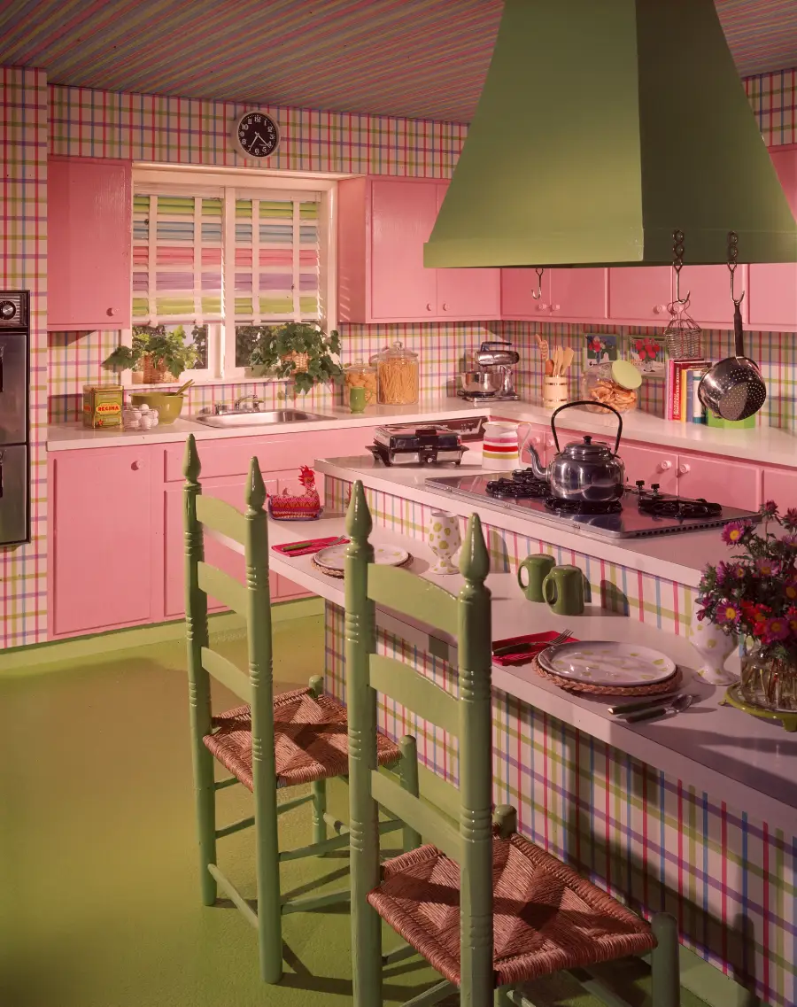 A retro kitchen with pink cabinets, green chairs, plaid wallpaper, green vent hood, and pastel decor. The counters hold dishes, a teapot, cookbooks, and plants, creating a bright, vintage atmosphere.