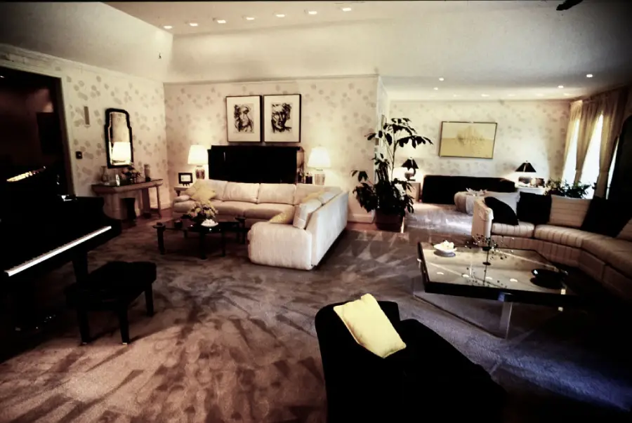 A spacious, elegant living room with cream sofas, a black grand piano, modern art on the walls, soft lighting, and large windows with sheer curtains. The decor is minimalist with plants and a glass coffee table.