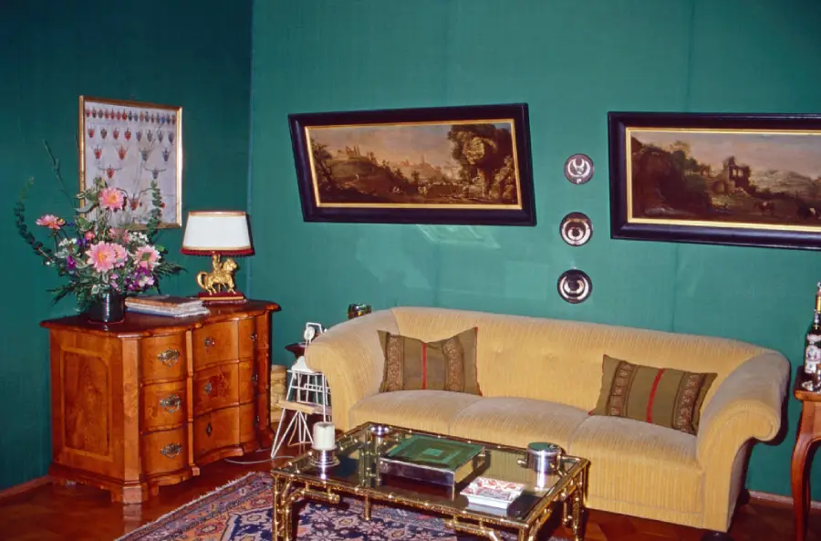 A living room with green walls, a yellow sofa, two crooked paintings, a wooden side table with flowers and a lamp, and a glass coffee table on a patterned rug.