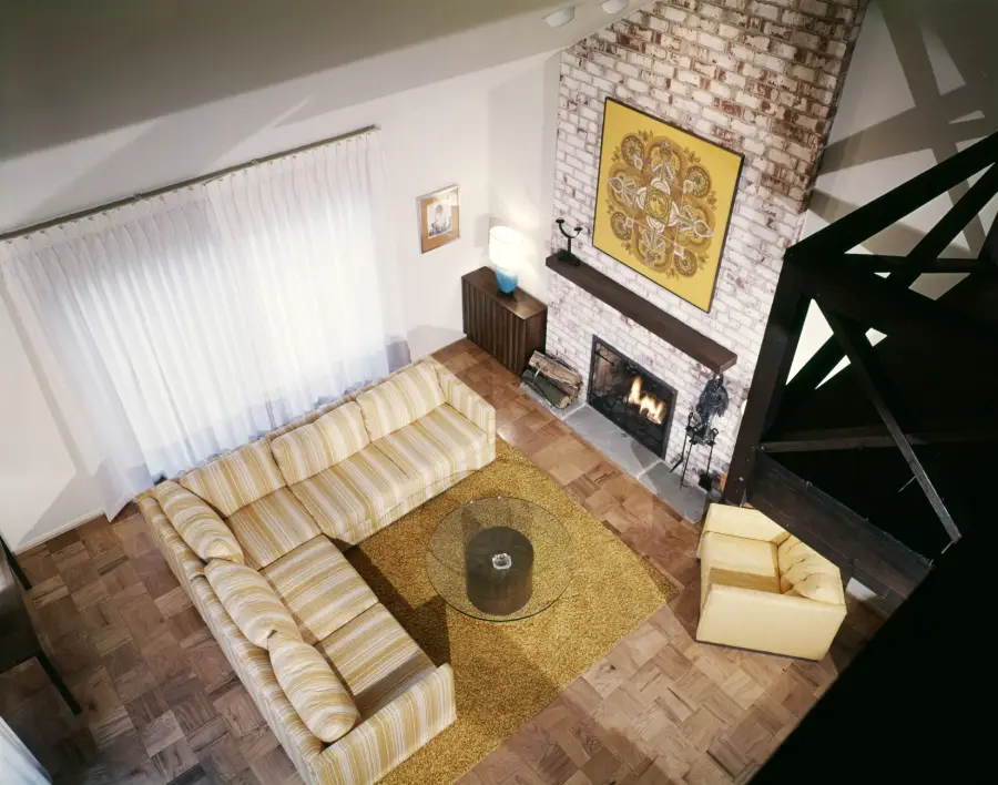 A mid-century modern living room with a striped yellow sectional, round glass coffee table, yellow armchair, shag rug, brick fireplace, and framed art above the mantel, viewed from above.