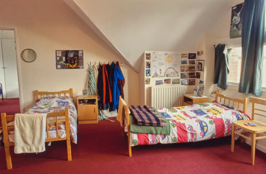 Two single beds with colorful bedding in a cozy room. Clothes hang on hooks, posters and photos decorate the wall, and sunlight comes through a window with green curtains. A small nightstand sits between the beds.