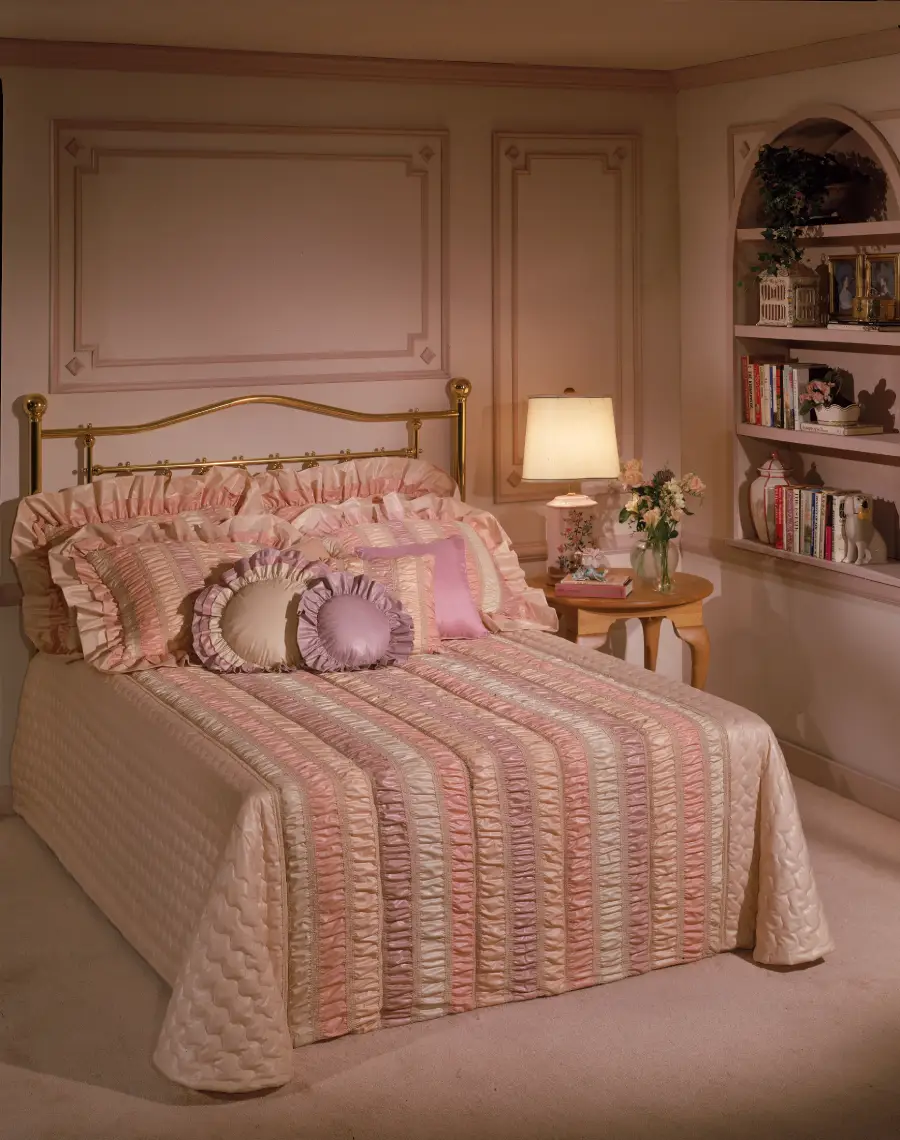 A neatly made bed with ruffled pillows and a pastel striped bedspread, next to a round table with flowers and a lamp, and a built-in bookshelf filled with books in a softly lit, elegant bedroom.