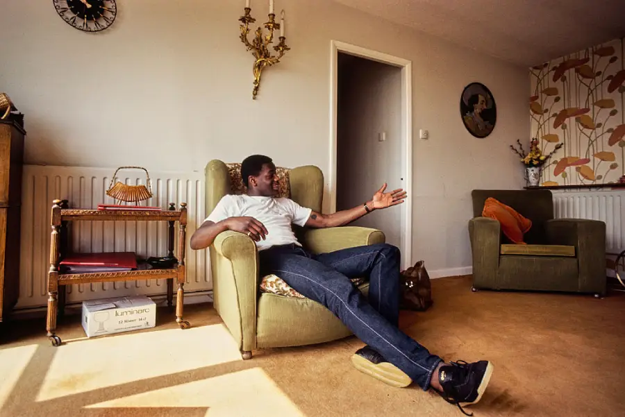 A man sits in an armchair in a living room, gesturing with his left hand. The room has vintage decor, with patterned wallpaper, wall clock, and a side table holding a lamp and basket.