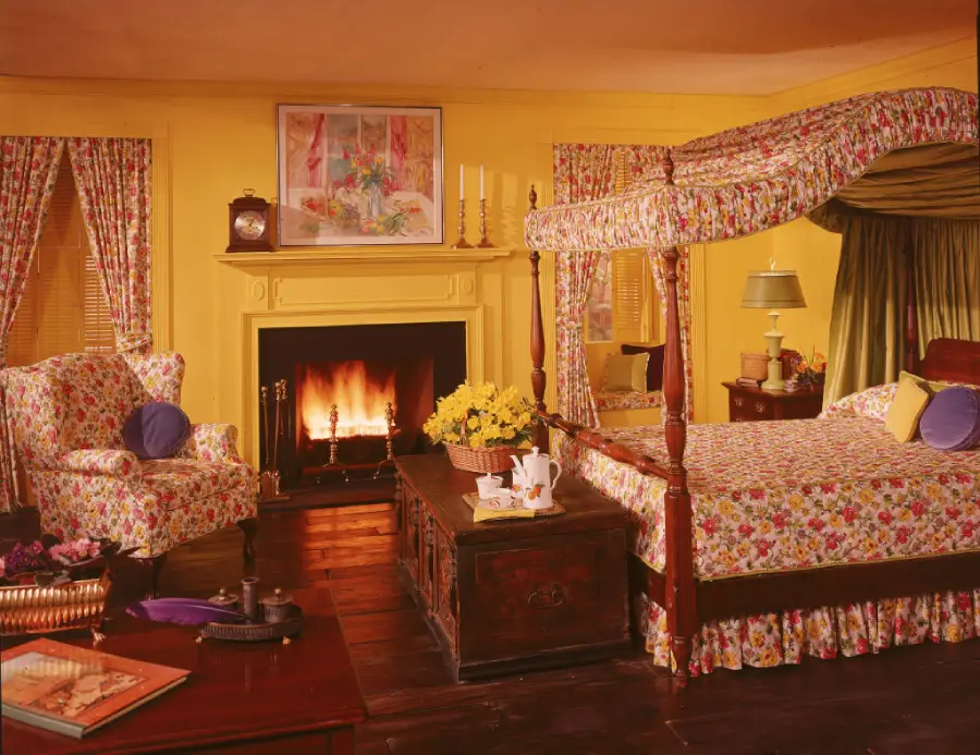 A cozy bedroom with yellow walls, a canopy bed, fireplace, floral-patterned bedding and curtains, armchair, wooden chest, and a coffee set on a tray. Flowers and framed art add charm.