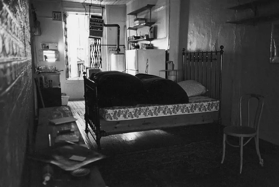 Black and white photo of a small, dimly lit room with a metal bed, floral quilt, chair, shelves, and a refrigerator in the background. Light enters through an open door at the far end.