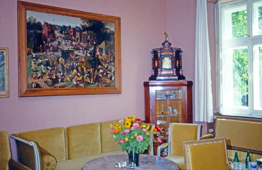 A cozy room with yellow couches, a marble table with a flower vase, a cabinet with figurines, a clock on top, a large painting on the wall, and a window with white curtains letting in natural light.