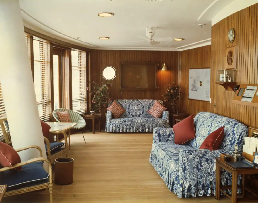 A cozy living room with wood-paneled walls, two blue patterned sofas with red pillows, rattan chairs, a small table, large windows with blinds, plants, and nautical decorations on the walls.