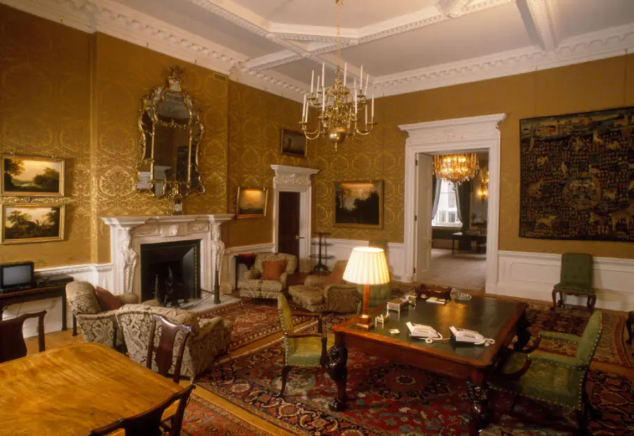Elegant, historic room with ornate gold wallpaper, chandelier, fireplace, antique furniture, large rugs, mirrors, paintings, and open doors leading to another equally grand space.