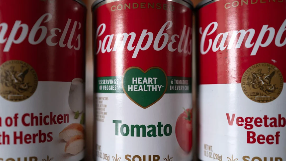 Three Campbell’s soup cans are shown close up. The center can is tomato flavor and features a green “Heart Healthy!” label. The other cans are partially visible: one is chicken with herbs, the other is vegetable beef.