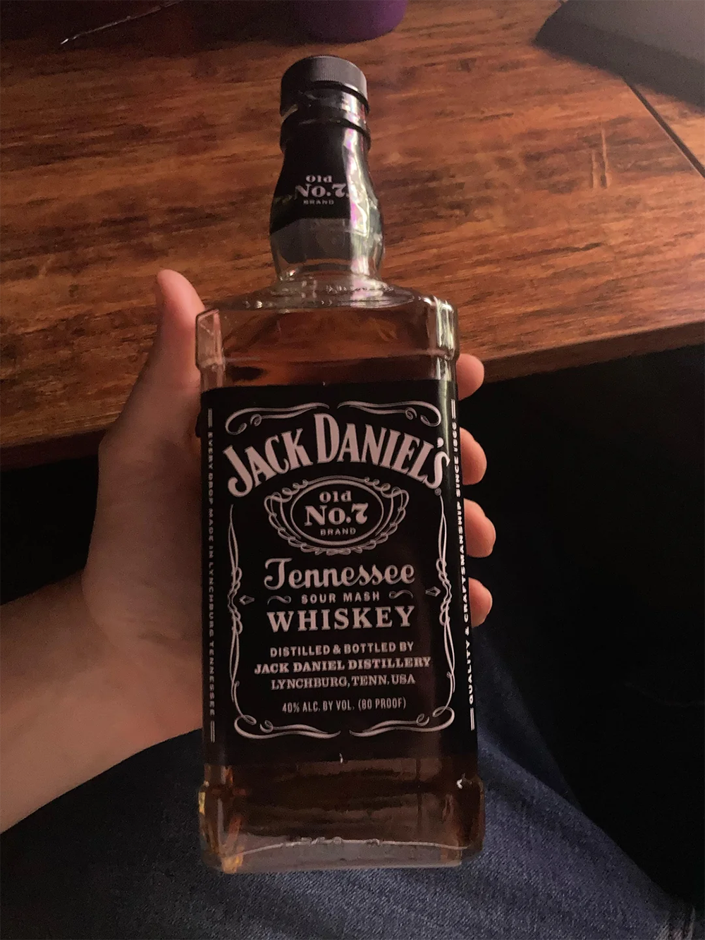A hand holding a bottle of Jack Daniel’s Old No. 7 Tennessee Sour Mash Whiskey over a dark wooden surface.
