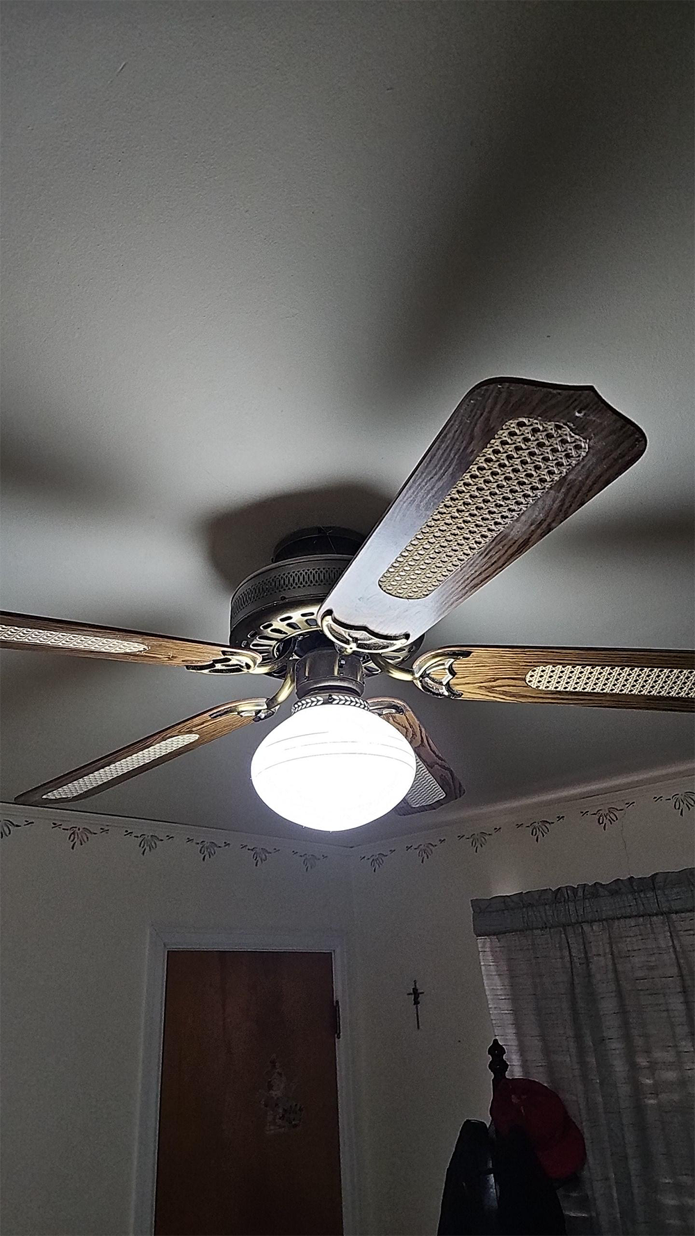 A ceiling fan with four wooden blades and a bright light fixture is mounted on a white ceiling. The room has a door, a window with curtains, and a small cross on the wall.