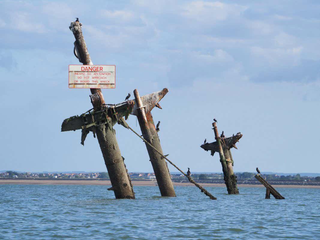 Rusty shipwreck remains protrude from the water, with birds perched on the metal beams. A sign warns “DANGER: Do not approach or touch wreck.” The distant shore and cloudy sky are visible in the background.