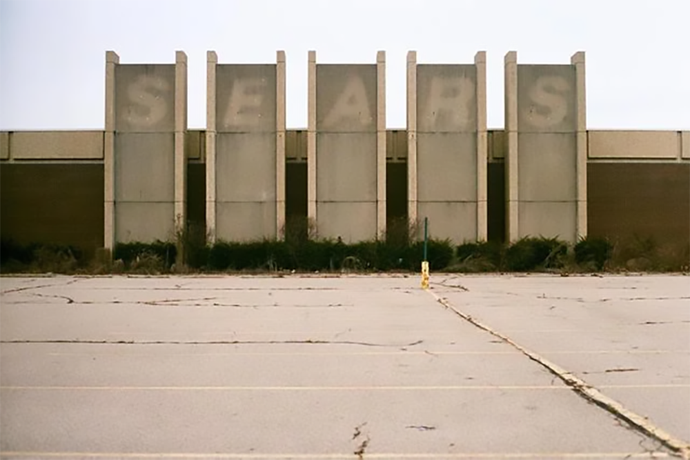 An abandoned, empty parking lot sits in front of a large, old building with faded “SEARS” letters visible on tall, rectangular panels. Shrubs grow along the building, adding to the scene’s neglected appearance.