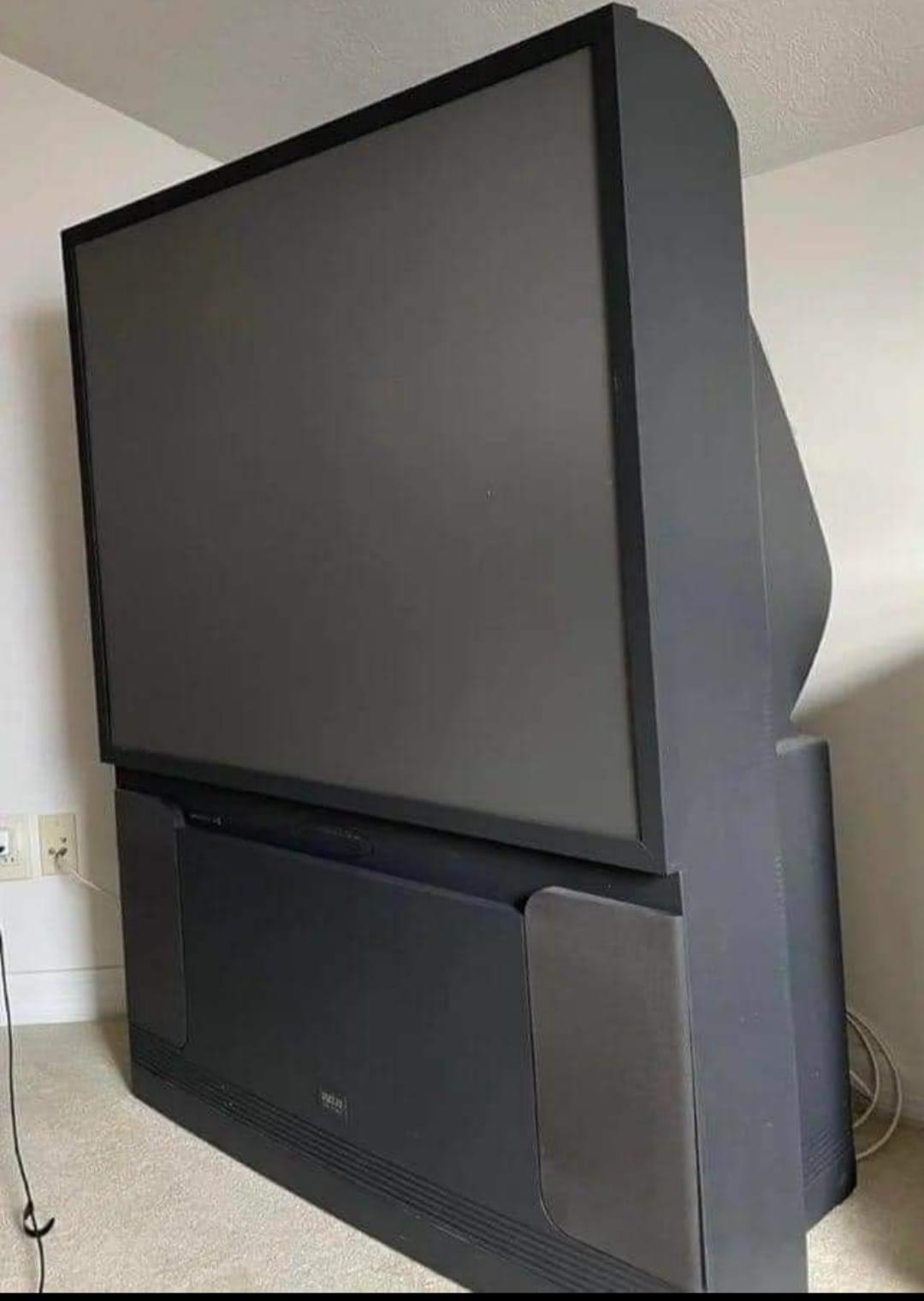 A large, old-fashioned rear projection television with a thick, deep design sits in the corner of a room on a beige carpet.