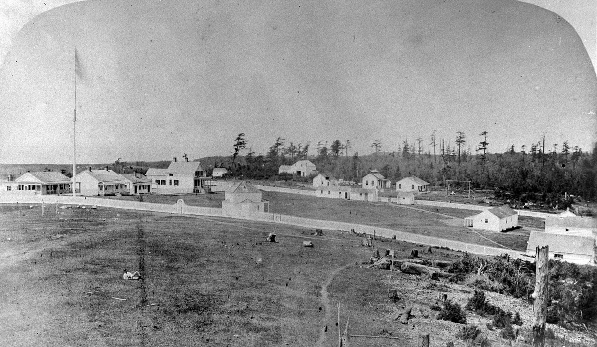 Black and white photo of a small rural settlement with scattered wooden buildings, dirt roads, open grassy areas, and trees in the background. The scene appears quiet and sparsely populated.
