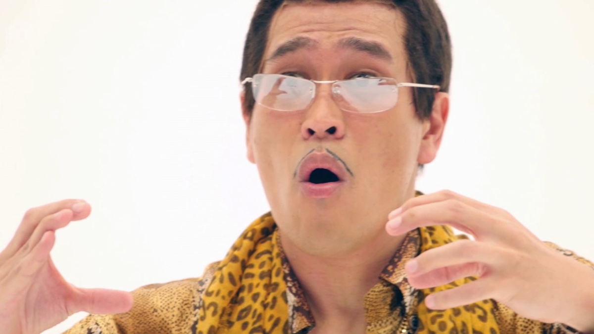 A man wearing glasses, a leopard print scarf, and a gold shirt gestures with his hands and has an expressive, surprised look on his face.