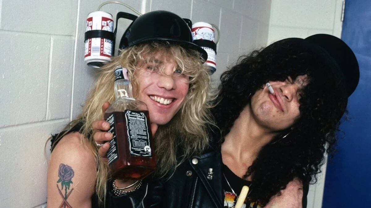 Two people with long hair pose together, smiling. One holds a bottle of Jack Daniel’s whiskey; the other has a cigarette in their mouth. Both wear black hats, and one has a beer can helmet. They’re dressed in rocker-style clothes.