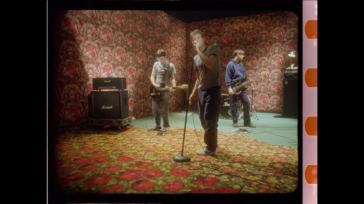 Three people play instruments in a band on a small stage with bold red-patterned wallpaper and a floral carpet. Song 2. One person sings into a microphone, while the others play guitar and bass. Music equipment is visible in the background.