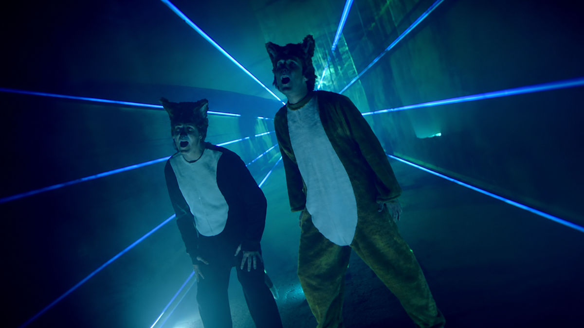 Two people in fox costumes stand in a dark room with blue laser lights crossing behind them, creating a dramatic atmosphere.