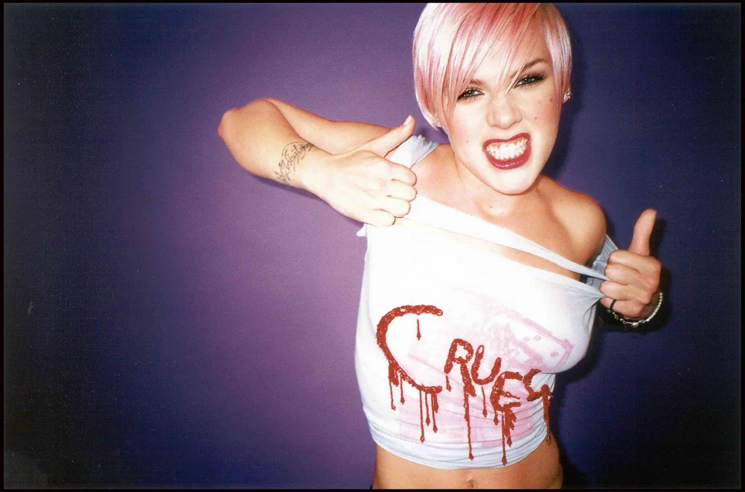Woman with short pink hair pulls at her white tank top, which has “CRUEL” written in red, dripping letters. She makes a fierce expression against a plain purple background.
