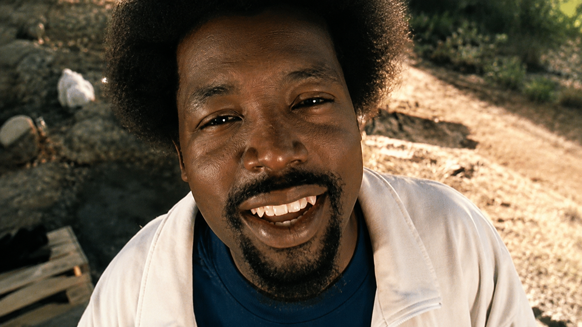 A man with an afro and goatee smiles warmly at the camera, wearing a white jacket over a blue shirt, with a sunlit outdoor background of dirt and rocks.