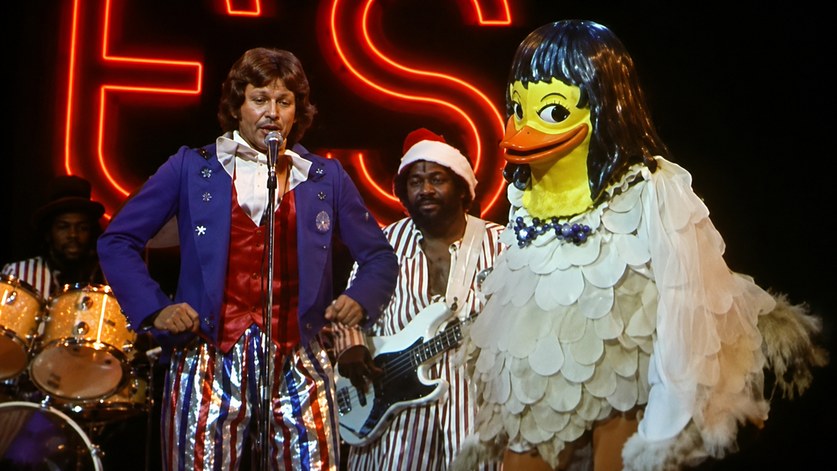 A man in a blue suit sings on stage next to a person in a large duck costume, with musicians playing instruments and a neon sign glowing in the background.