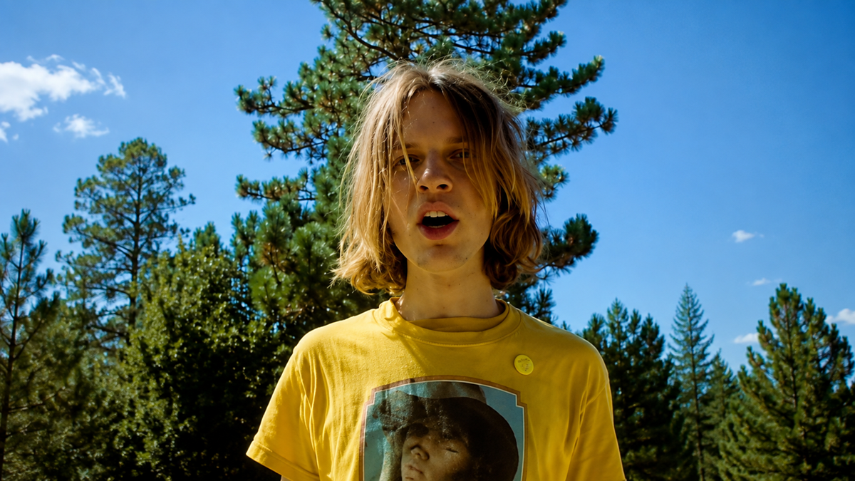 A person with shoulder-length blond hair wearing a yellow T-shirt stands outdoors in front of tall pine trees under a clear blue sky.