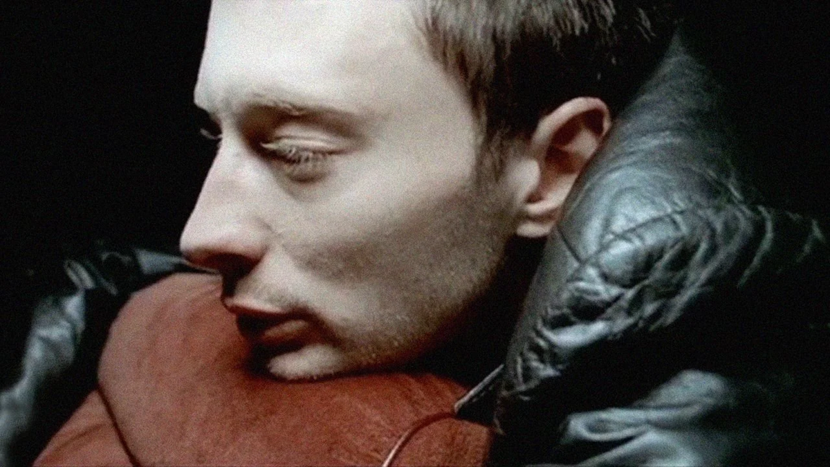 A person with short hair rests their head on a leather surface, looking thoughtful and pensive. The lighting is dim, highlighting the side of their face and the textures of the leather and clothing.