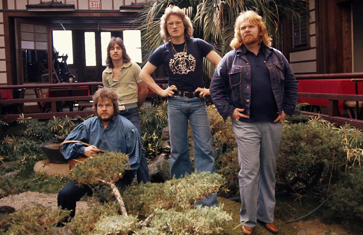 Four men with 1970s hairstyles and clothing pose indoors by a small garden with bushes; three are standing, one is crouched. The background has wood paneling, plants, and a glass door.
