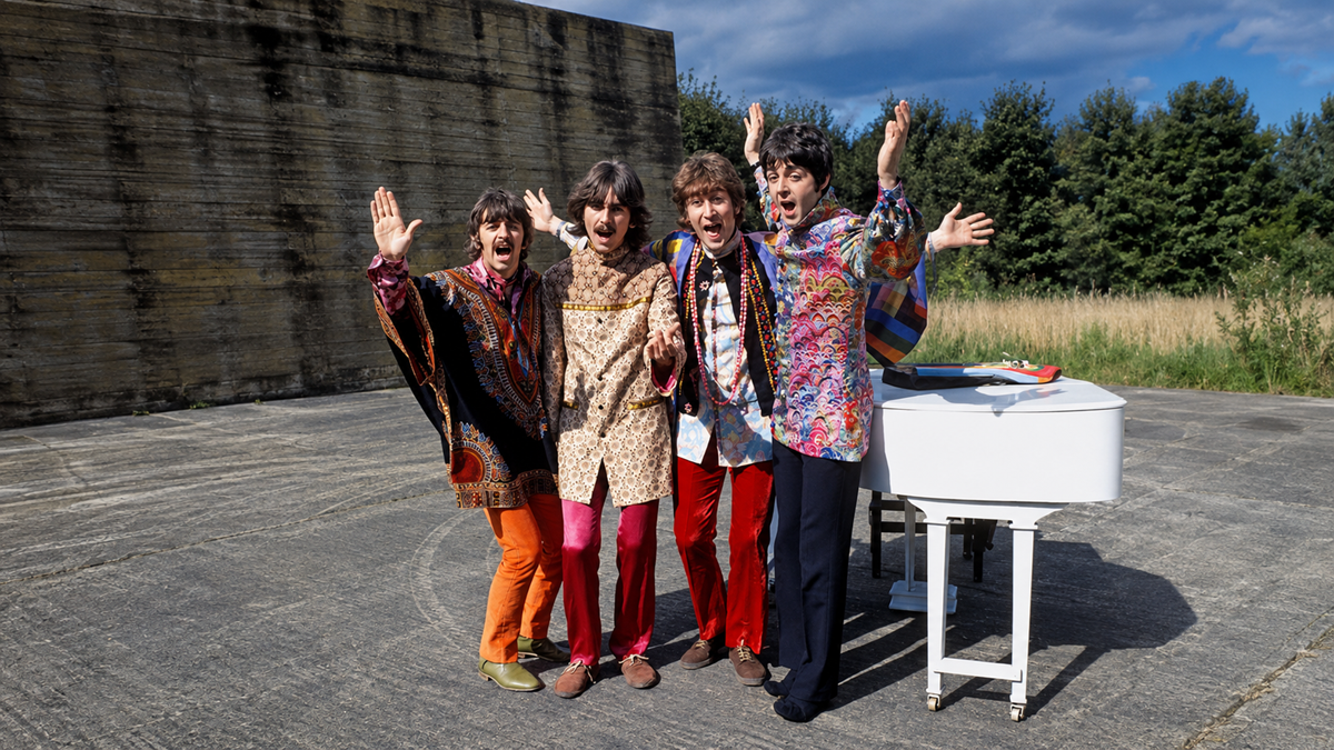 Four people in colorful, retro clothing stand on a concrete surface with a white piano, smiling and waving in front of a wall and greenery under a partly cloudy sky.