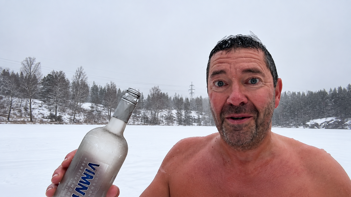 A man with wet hair stands shirtless in a snowy outdoor setting, holding a frosted bottle and looking surprised or amused. Snow-covered trees and cloudy sky are in the background.