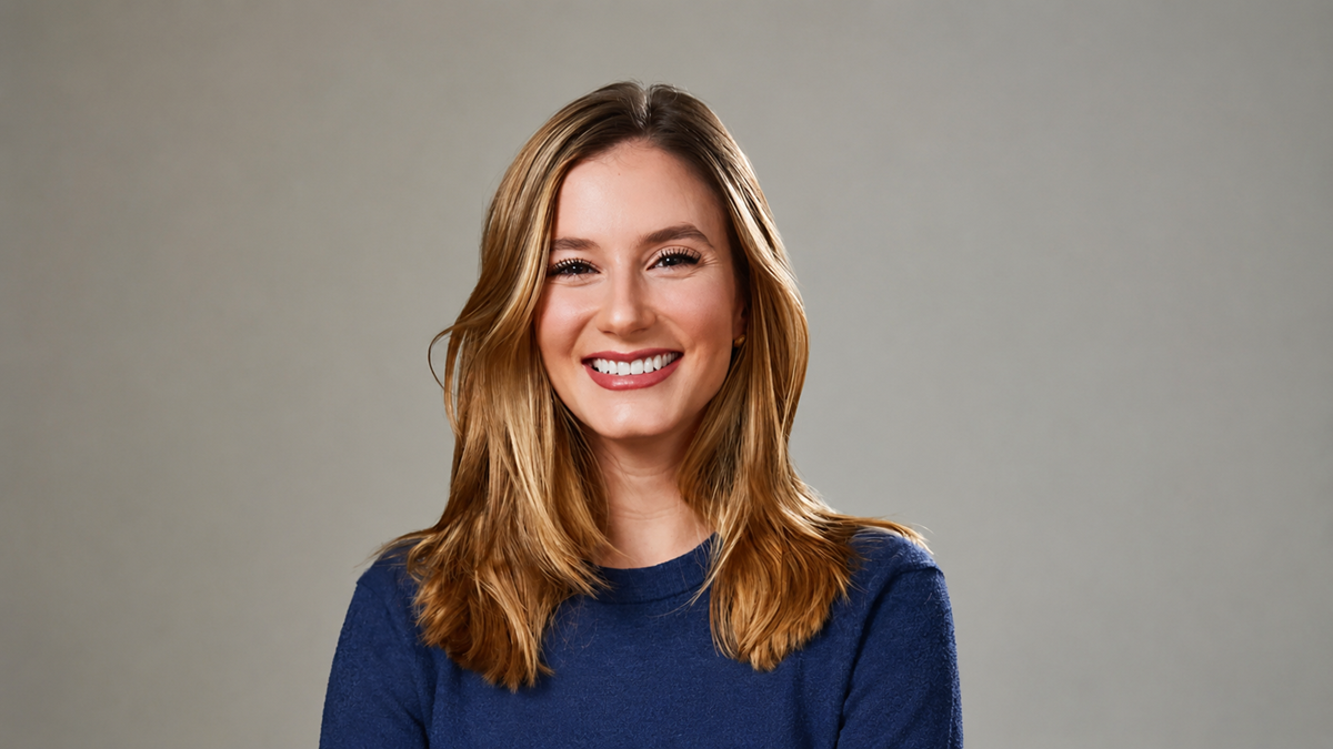 A woman with long, light brown hair wearing a blue sweater is smiling in front of a plain, light gray background.