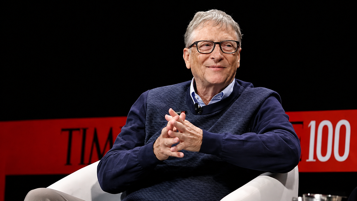An older man with glasses and gray hair, wearing a navy sweater over a collared shirt, smiles while sitting in a white chair on stage with a red and black “TIME 100” backdrop.
