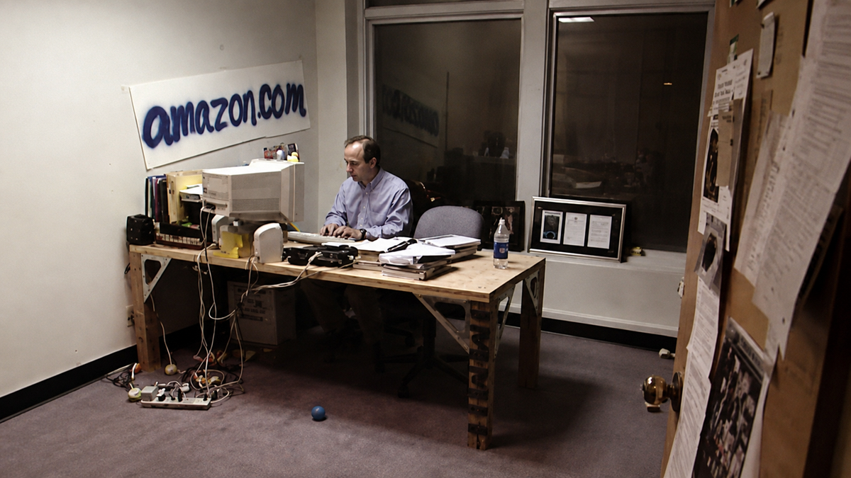A man works at a cluttered desk with an old computer in a sparsely furnished office. A hand-painted “amazon.com” sign hangs on the wall behind him.