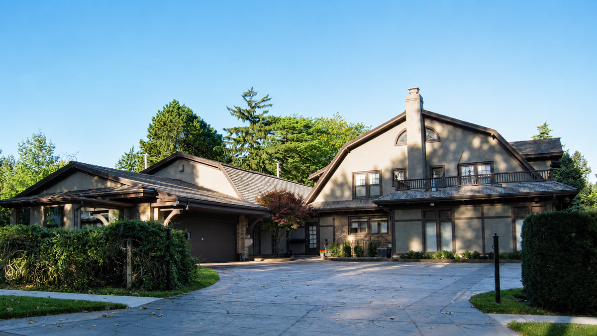 A large, two-story suburban house with tan walls, a dark roof, an attached garage, and a spacious driveway. Trees and shrubs surround the property under a clear blue sky.