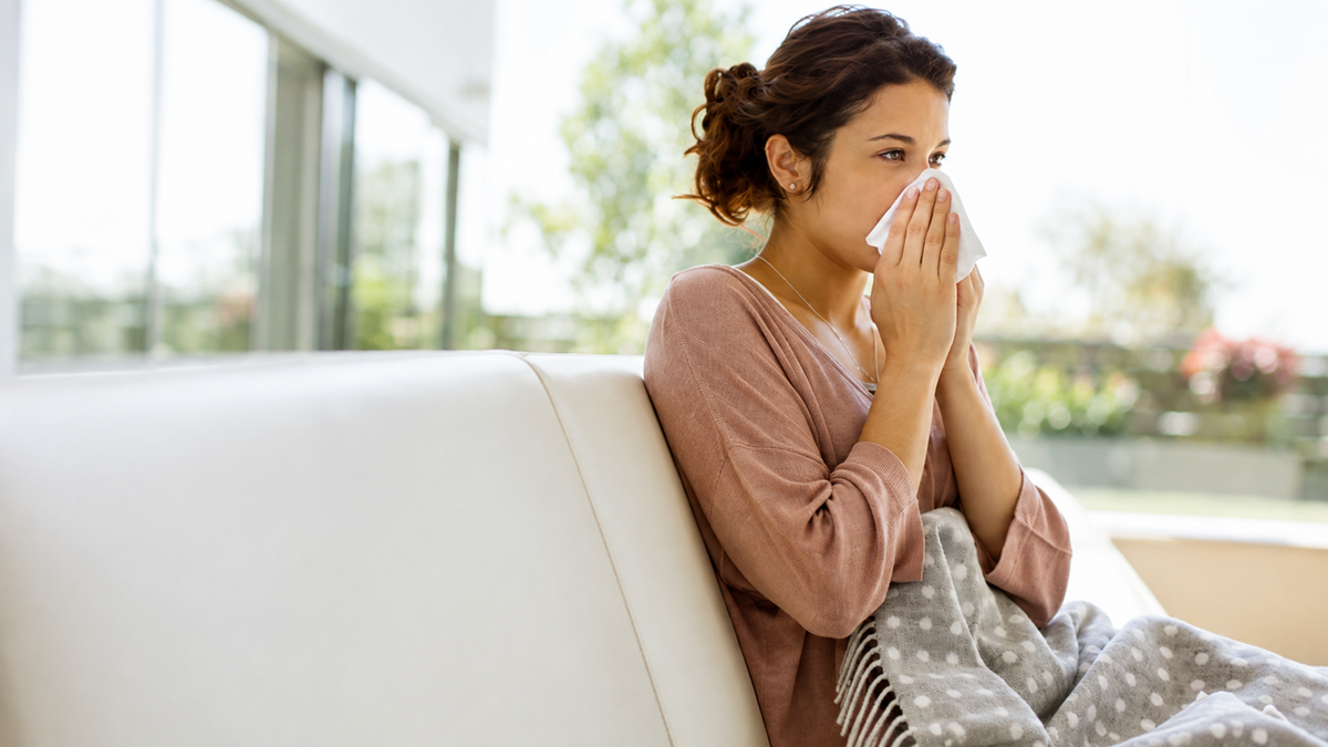A woman sitting on a couch, wrapped in a gray polka dot blanket, holds a tissue to her nose, appearing to sneeze or blow her nose. Sunlight streams through windows in the background.