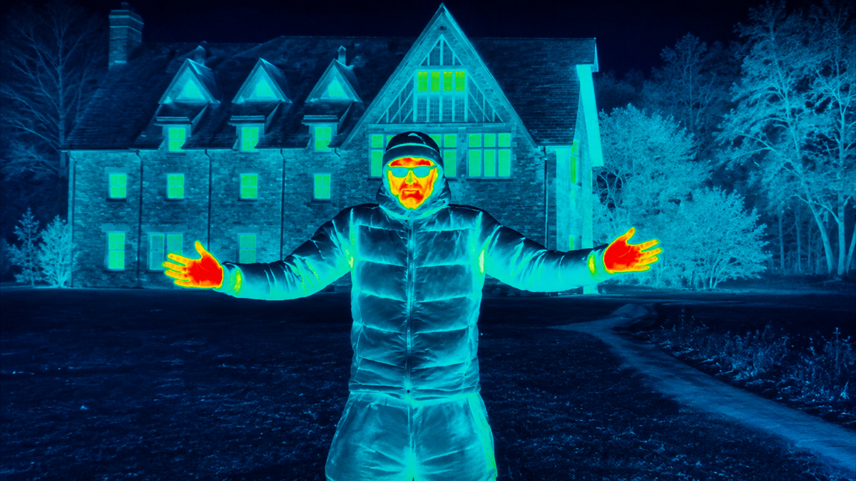 A thermal image of a person standing outside with arms outstretched in front of a large house at night. Warmer areas on the person’s body appear in red and yellow; the background is mostly blue and green.