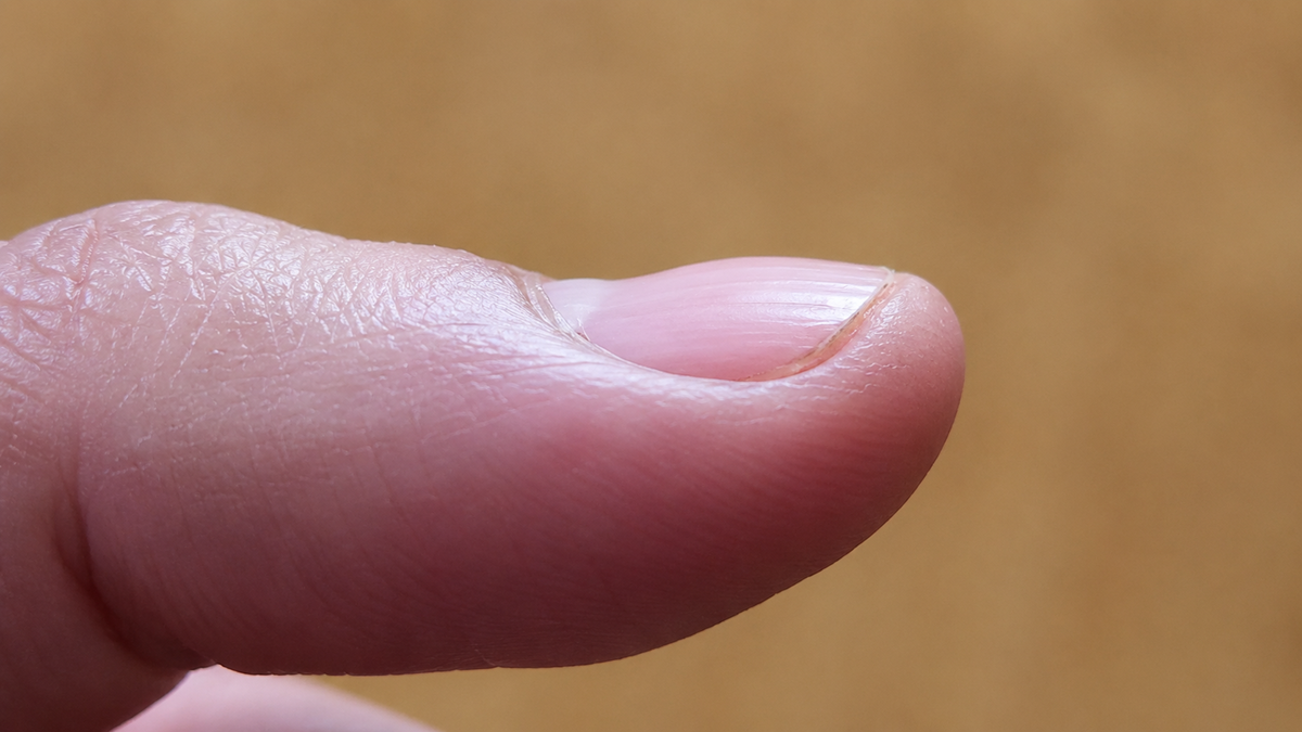 A close-up side view of a person’s thumb with a short, clean fingernail, set against a blurred brown background.