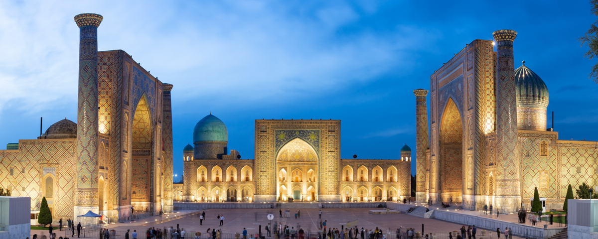 Evening view of Registan Square in Samarkand, Uzbekistan, featuring three grand madrasahs with intricate tilework, large arched entrances, and blue domes, illuminated against a deep blue sky.
