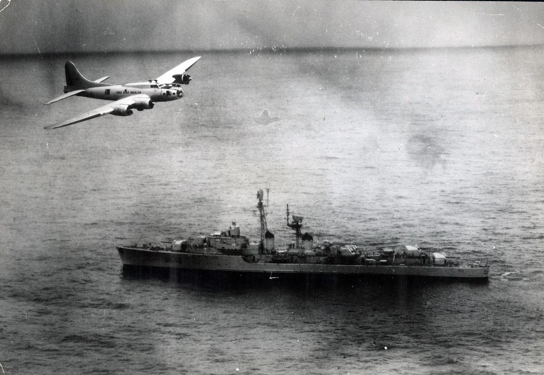 A military aircraft flies over a naval warship at sea. The black and white image shows the plane in mid-air above the ship, which is surrounded by calm waters.