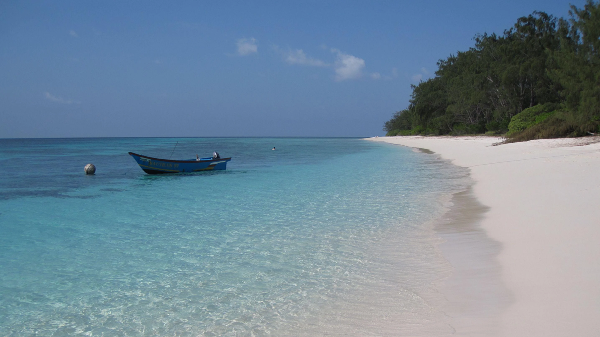 A blue boat floats on clear turquoise water near a white sandy beach with green trees under a blue sky.