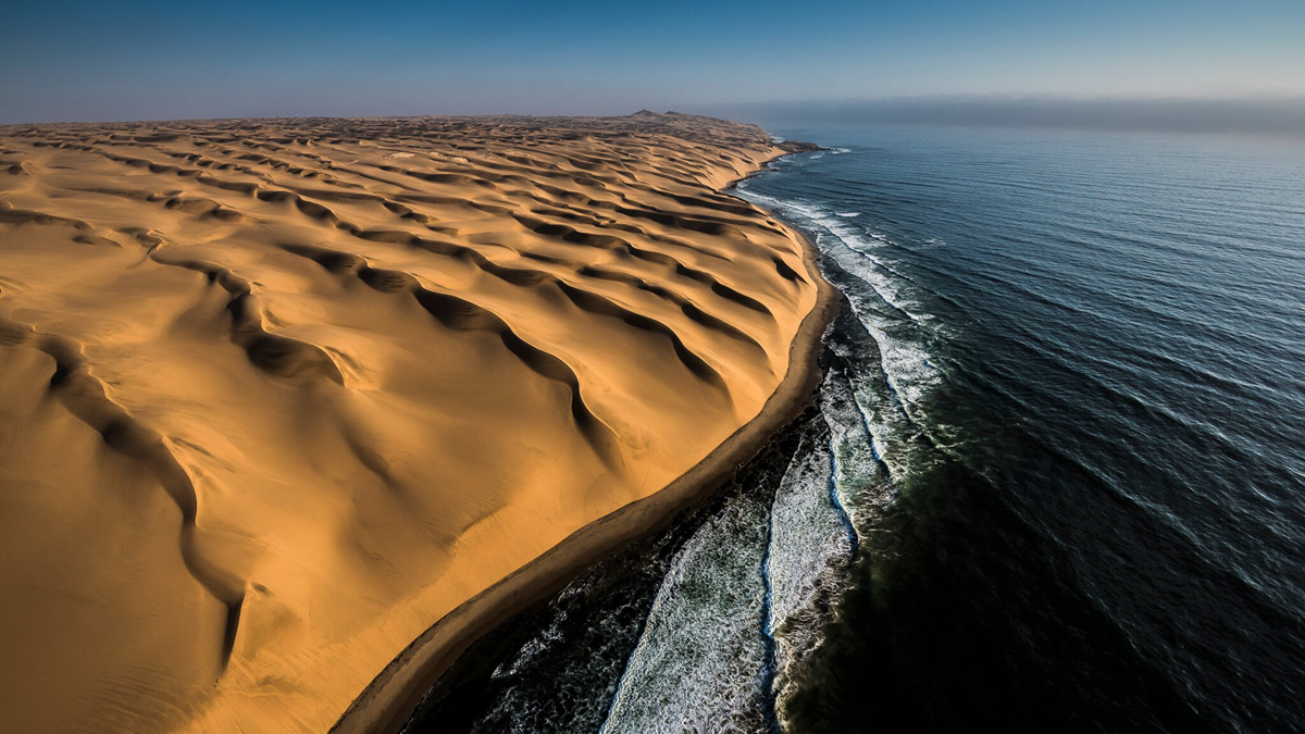 Aerial view of golden sand dunes meeting the dark blue ocean, with waves breaking along the shoreline under a clear sky.