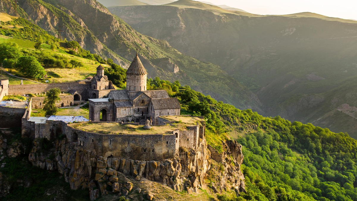 Ancient stone monastery perched on a rocky cliff overlooking a deep green valley and mountains under a golden sunset.