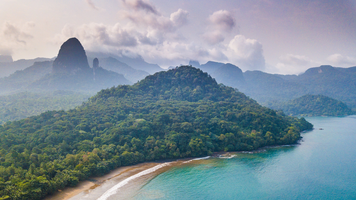 Aerial view of a lush, green tropical island with mountains in the background, a sandy beach, and turquoise ocean water under a partly cloudy sky.