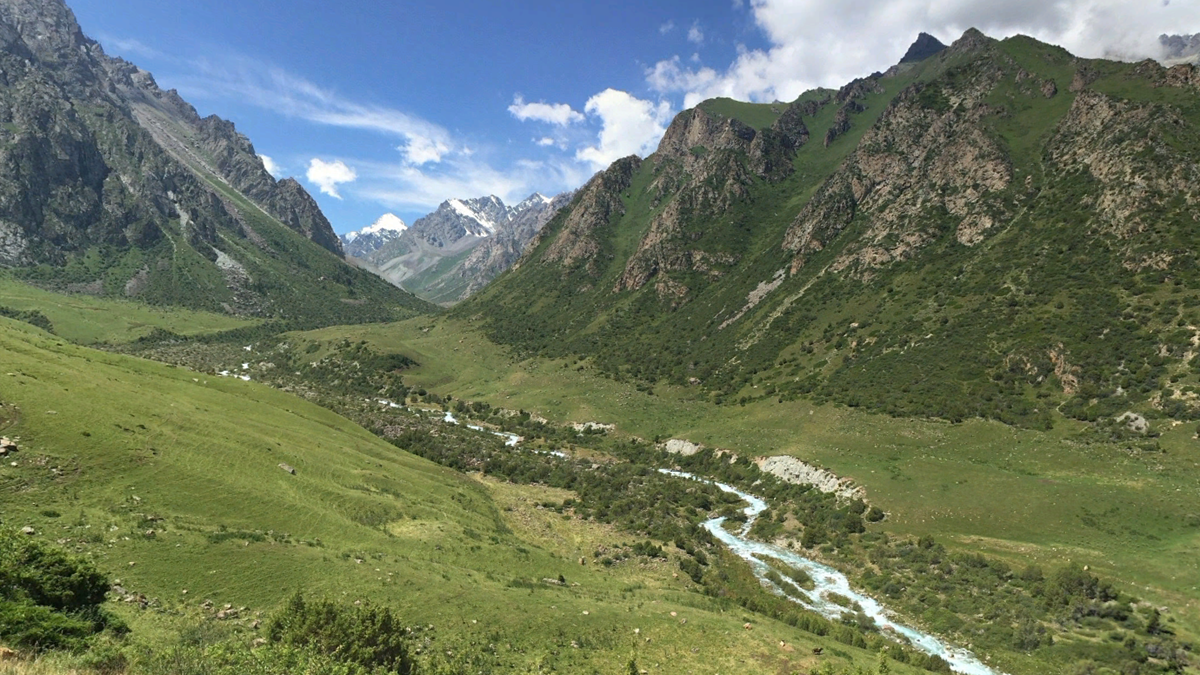 A lush green valley with a winding river is surrounded by tall, rocky mountains under a blue sky with scattered clouds. Snow-capped peaks are visible in the distance.