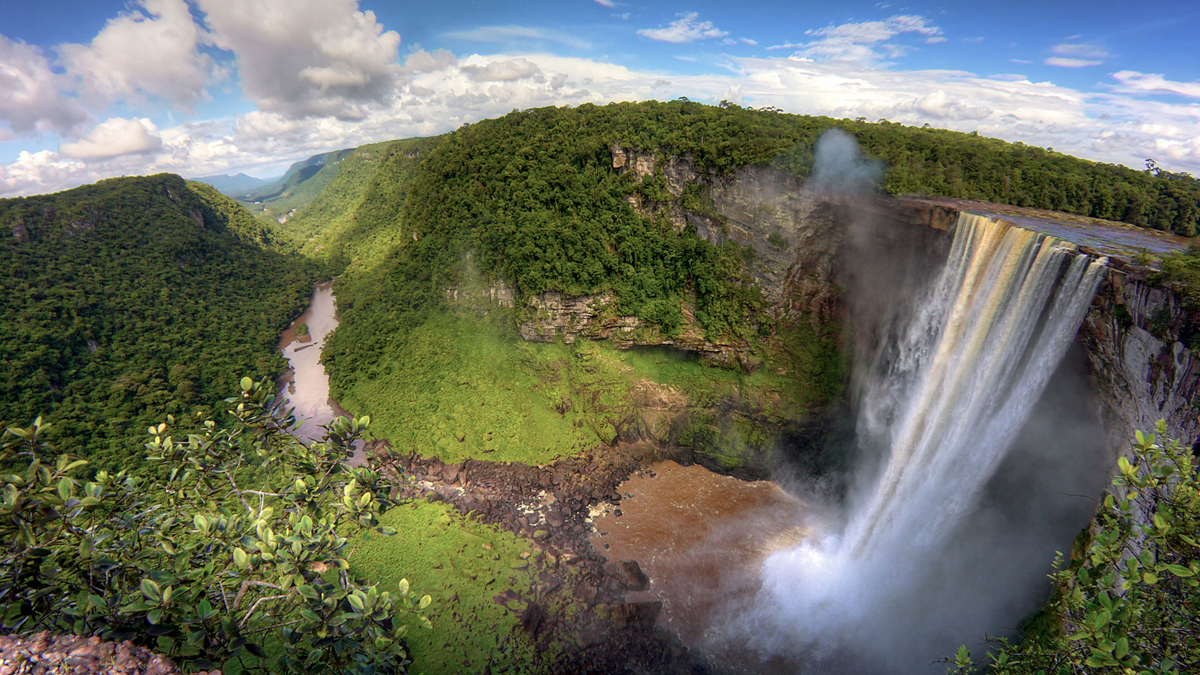 A large waterfall cascades over a cliff surrounded by lush green forest, with a river winding through a deep valley under a partly cloudy blue sky.