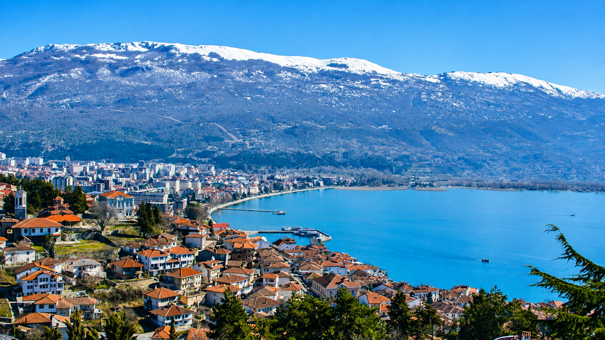 A panoramic view of a lake with a town of red-roofed buildings in the foreground, surrounded by hills and snow-capped mountains under a clear blue sky.