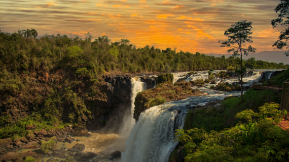 A lush green landscape with a powerful waterfall flowing into a rocky river, surrounded by dense trees, under a vibrant orange sunset sky.