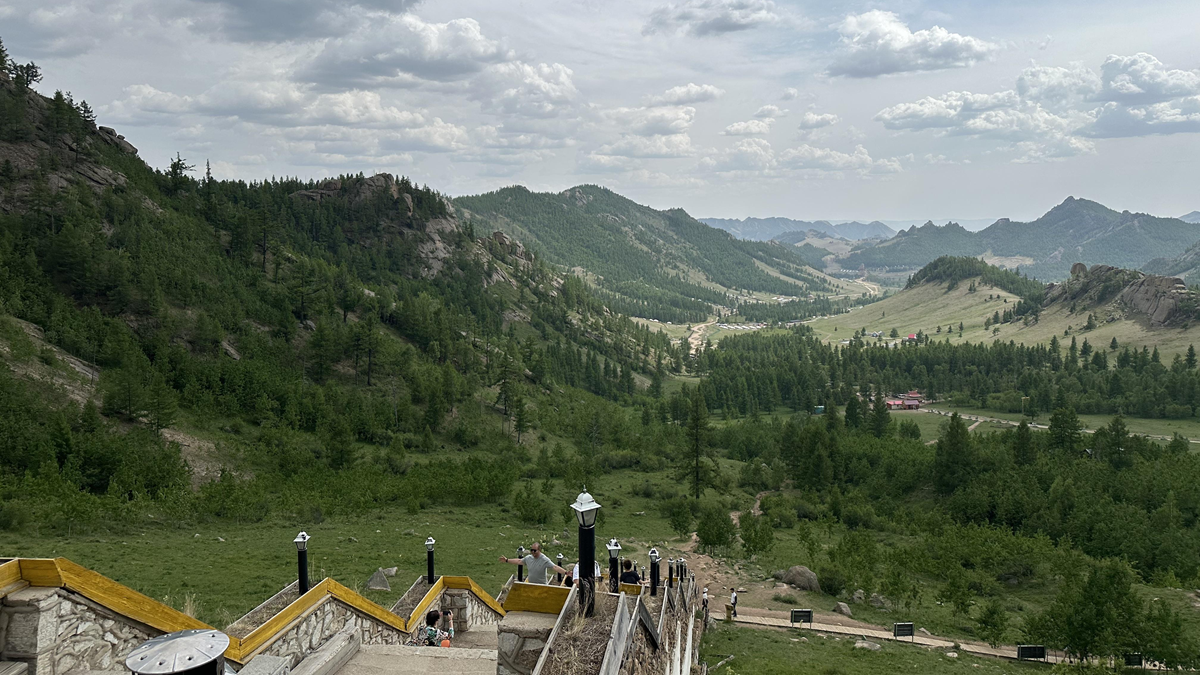 View from a stone staircase overlooking a lush green valley with hills, forests, and distant mountains under a cloudy sky. Several people stand near the stairs, enjoying the scenic landscape.