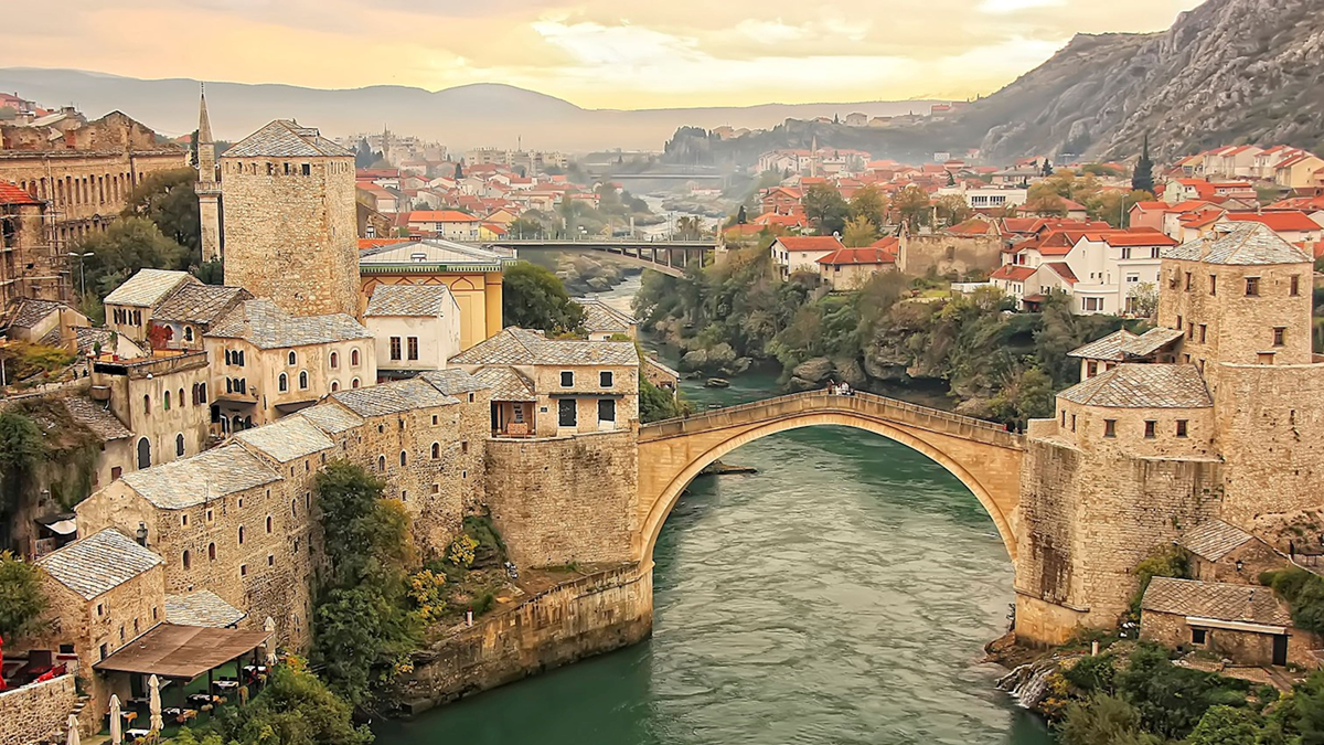 A stone arch bridge crosses a river, surrounded by historic buildings with tiled roofs in a picturesque town set against hills at sunset. countries